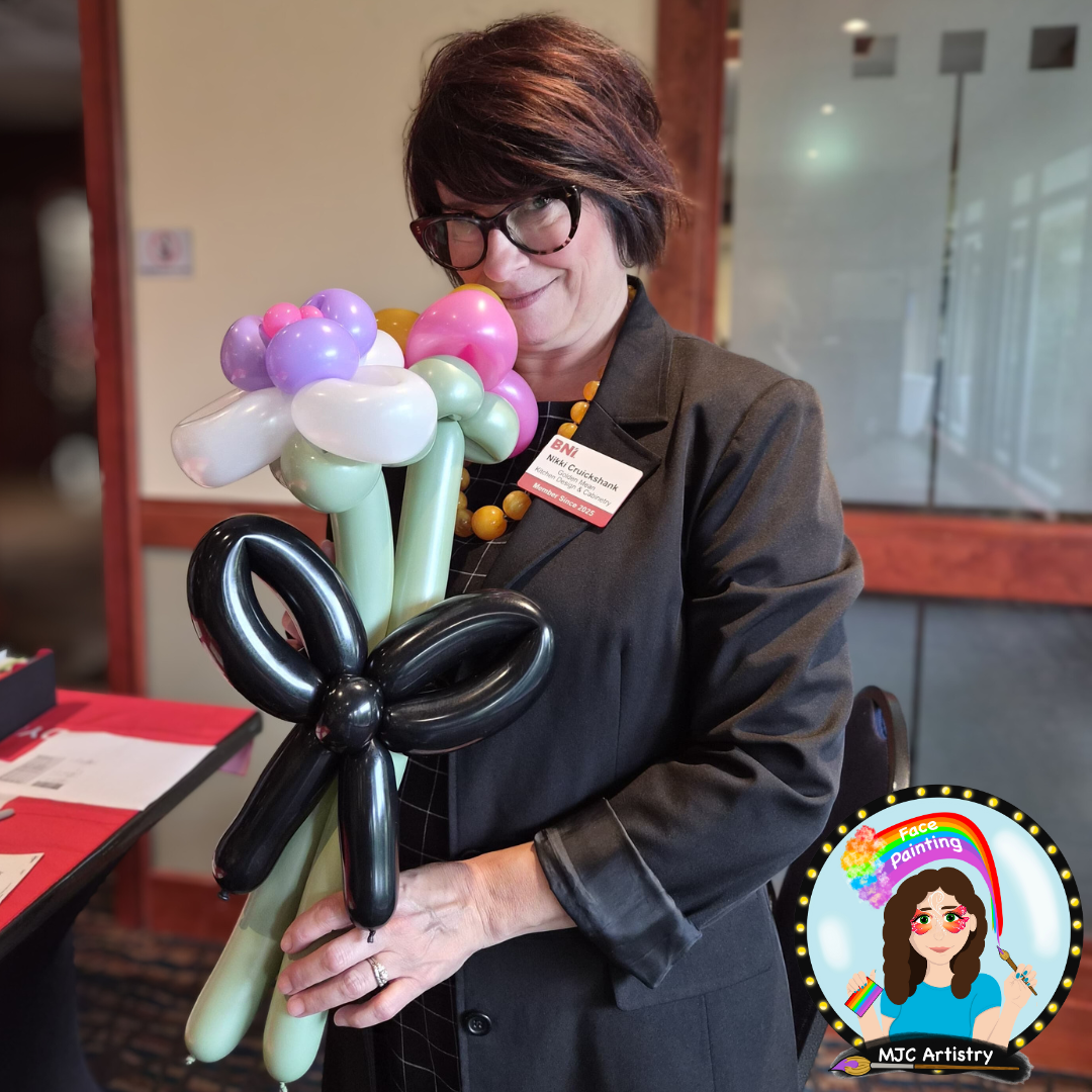 Woman with short dark hair, glasses, and a black blazer holding a balloon bouquet resembling a flower. She is wearing a name tag and a colorful beaded necklace, standing indoors with a table and glass wall in the background.