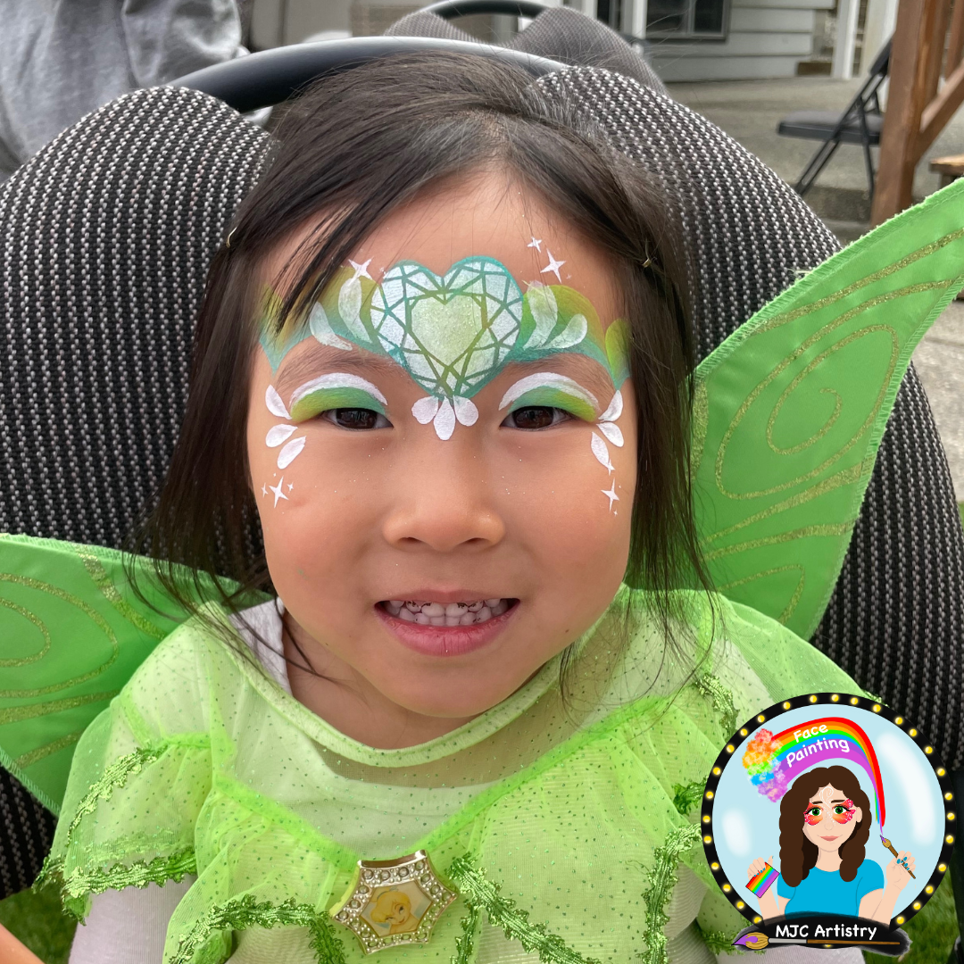 Young girl dressed as a fairy with green wings, face paint featuring a green heart, white and green accents, and a costume with a Disney princess badge. She is smiling at the camera, sitting on a black and white patterned chair outdoors.