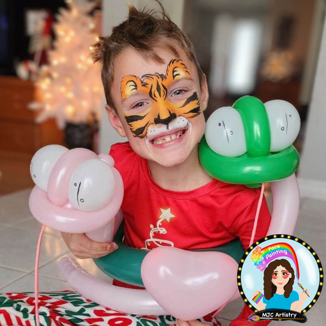 A young boy with face paint resembling a tiger, smiling widely, holding balloons shaped like hearts and carrying a balloon wreath, celebrating Christmas indoors.