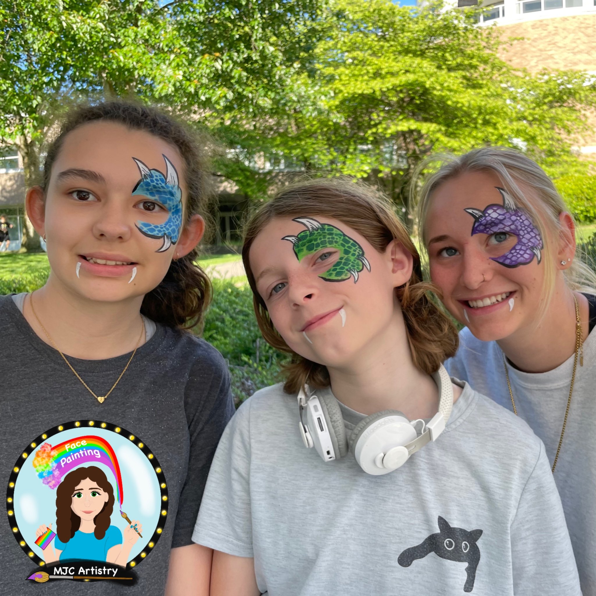 Three teens with colorful dragon face paint, two girls and one boy, smiling outdoors in a park with trees and grass.