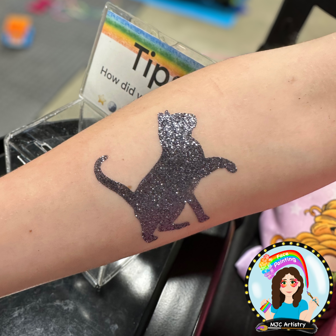 Glitter tattoo of a cat on a person’s arm with a tip card in the background at a face painting station in Vancouver.