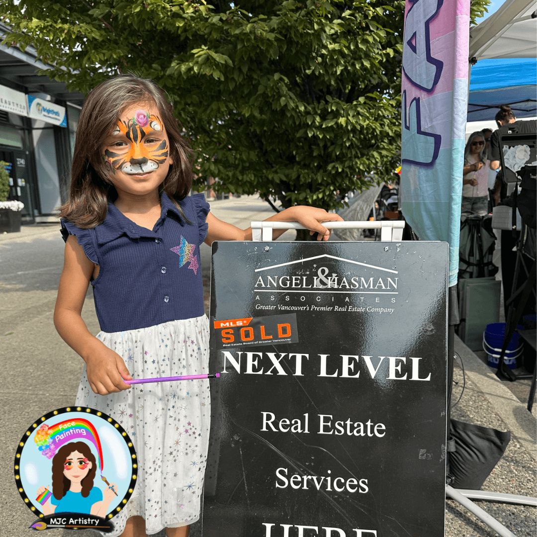 A young girl with face paint resembling a tiger, smiling and holding a stick, standing next to a sign for a real estate company at an outdoor event.