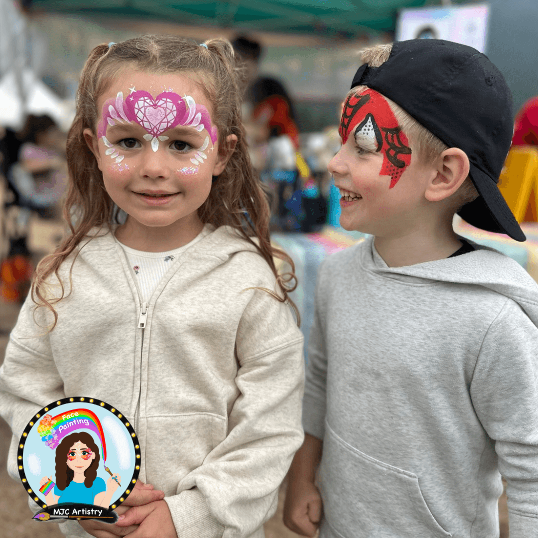 Two children with face paint, smiling at an outdoor event. The girl has pink and white butterfly face paint, and the boy has red and black spider web face painting in Vancouver BC.