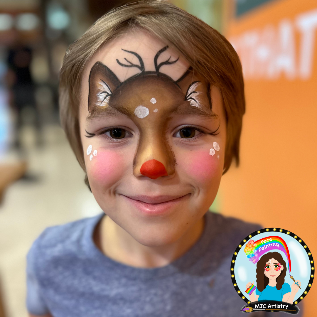 Young boy with face paint resembling a reindeer, including antlers, a brown face, a red nose, and white accents, smiling at the camera at an event in Vancouver. 