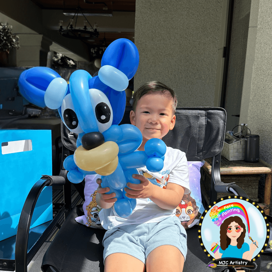 A young boy sitting in a black outdoor chair, smiling and holding a balloon sculpture of a cartoon dog. The boy is wearing a white t-shirt and gray shorts, with a decorative face painting pillow behind him taken at a birthday party in Vancouver BC. 