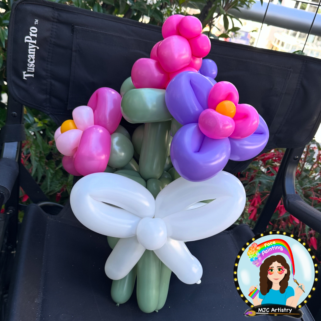 Colorful balloon bouquet arranged to resemble a flower with purple, pink, green, and white balloons, set on a black chair with a logo of a face painter and rainbow in the bottom right corner.