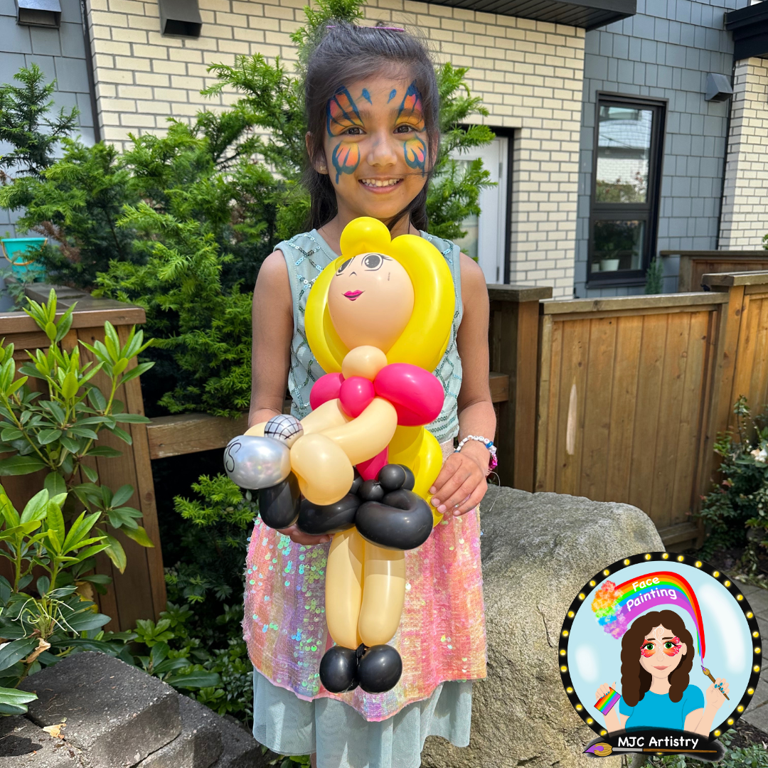 Young girl with butterfly face paint smiling and holding a balloon sculpture of a princess with yellow hair, pink and beige dress, and black shoes outdoors near a brick house, greenery, and wooden fencing.