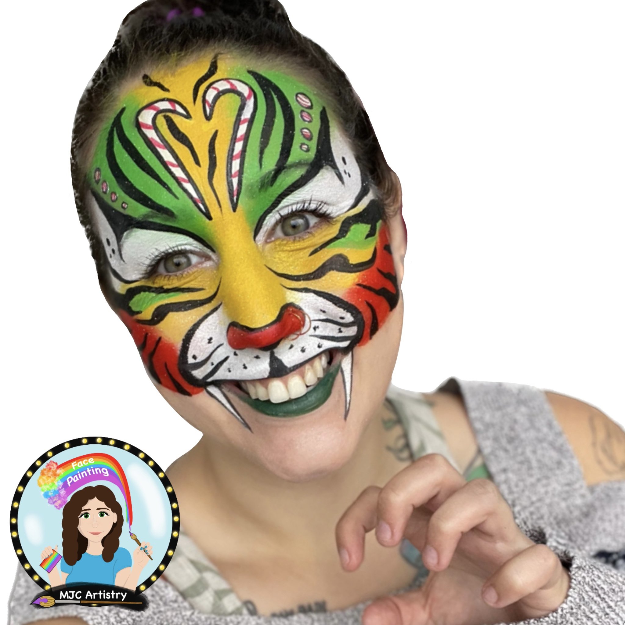 Woman with colorful face paint resembling a tiger, smiling and holding her hand near her face, with a tattoo on her arm and a gray top. Logo in bottom left corner reads "Face Painting" by MJC Artistry.