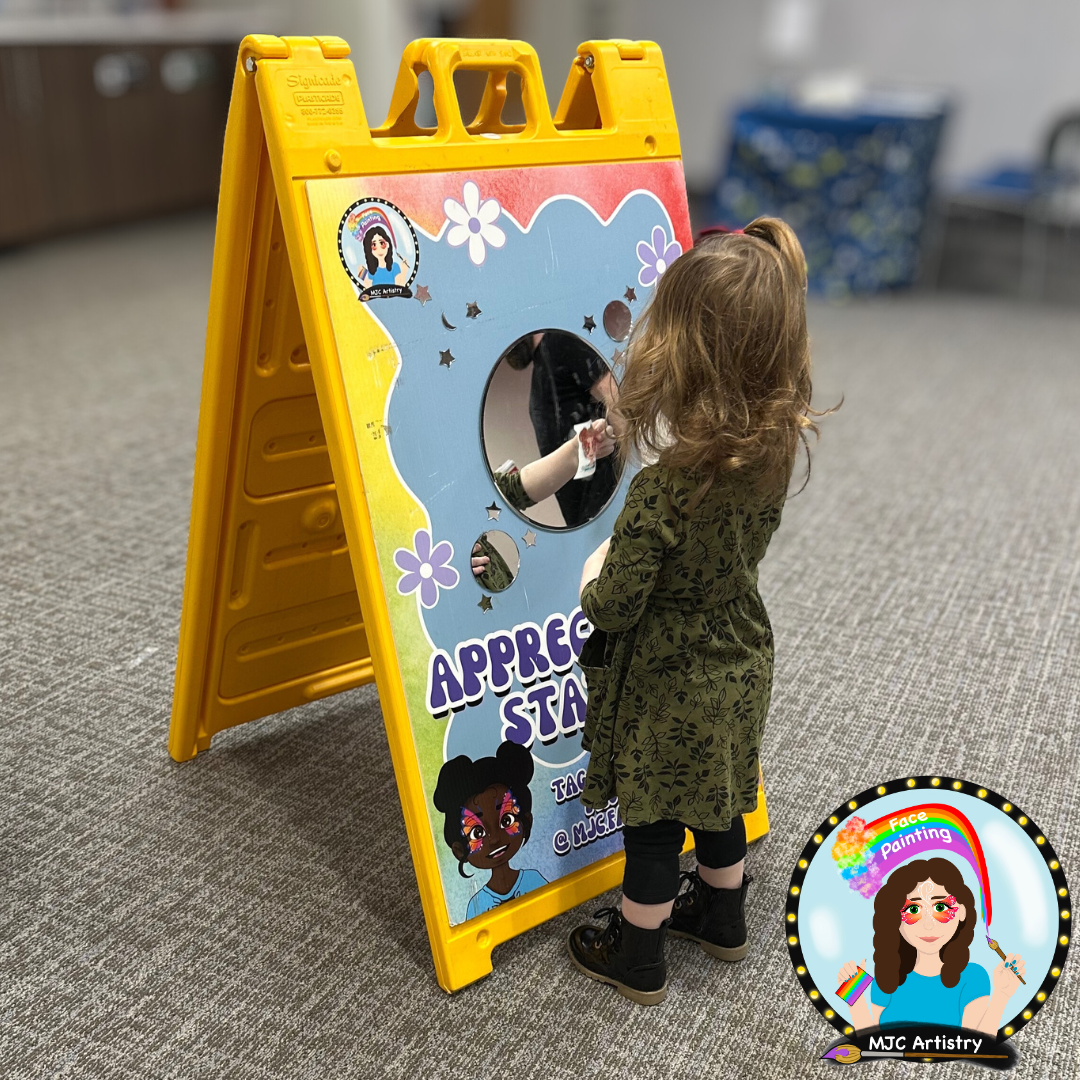 A young girl with curly hair wearing a green dress and black boots, standing in front of a colorful appreciation stand with a mirror, part of face painting activity at a children's event in Vancouver BC