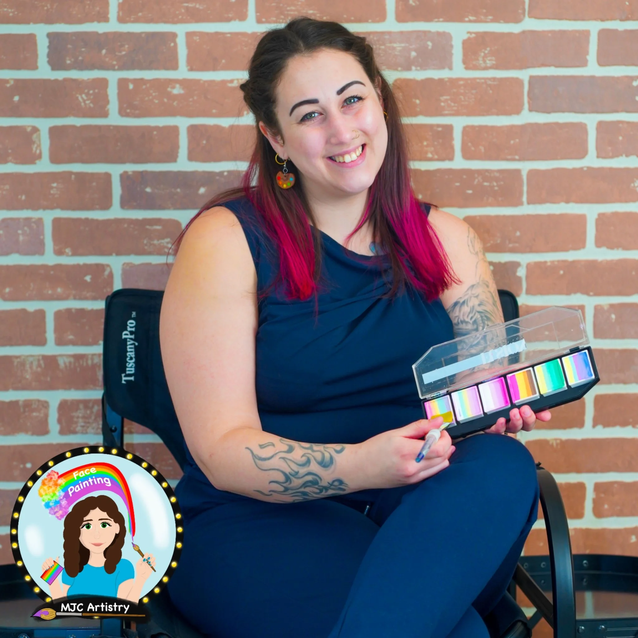 Owner and face painter of MJC Artistry with pink hair, tattoos, and colorful earrings smiling while holding a box of face paint colors and a brush, seated in front of a brick wall posing for a photo for her face painting business in Vancouver BC