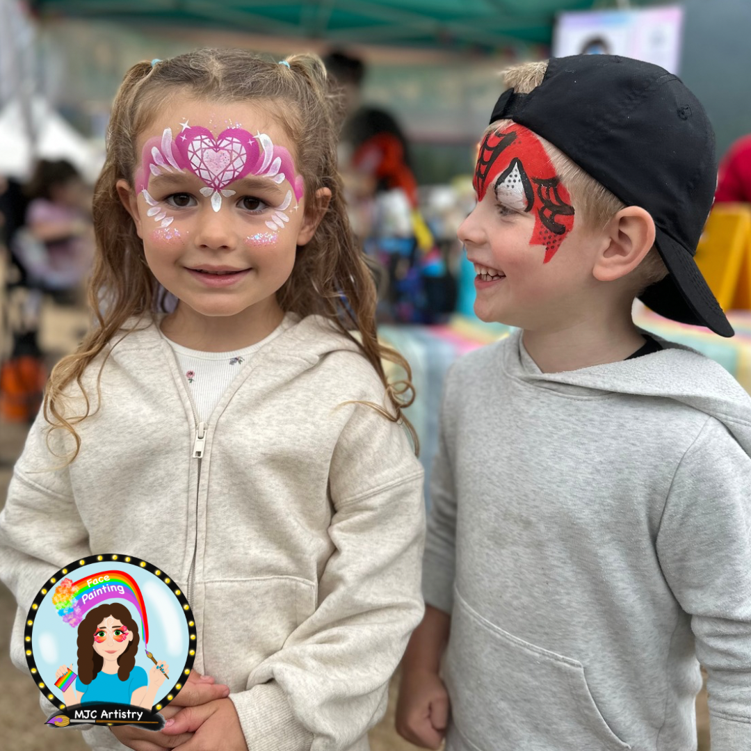 Young girl with face paint of a pink heart with white wings and sparkles, smiling at camera, standing next to a boy with face paint of spider man, wearing a black cap backward, at an outdoor event in Vancouver BC.