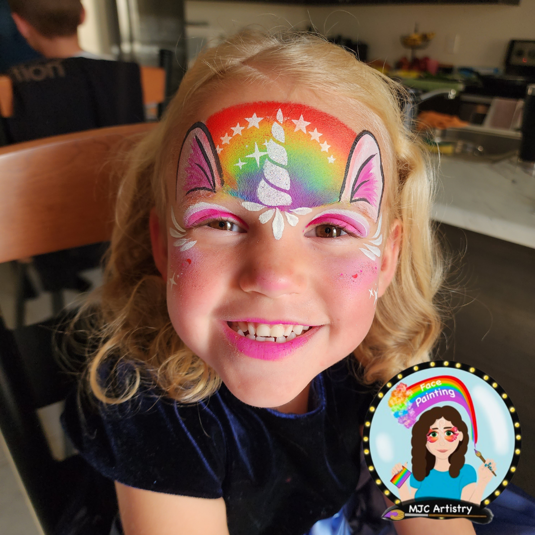 Little girl smiling with colorful face paint resembling a unicorn and rainbow, with star accents. Background shows a kitchen and dining area.