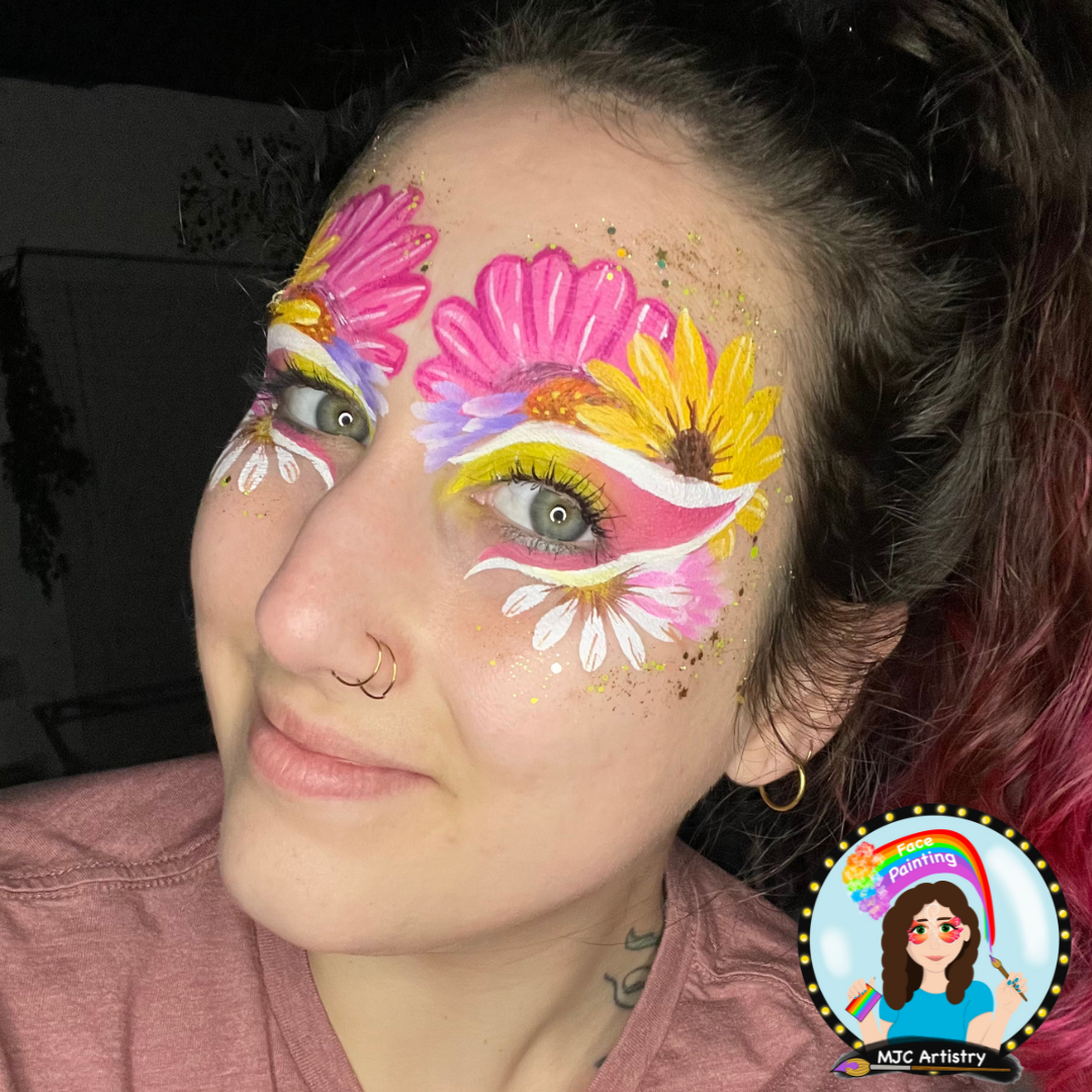 A woman with face paint resembling colorful flowers around her eyes, featuring pink, yellow, white, and purple hues, with some glitter accents. She has multiple facial piercings and is wearing a pinkish t-shirt.