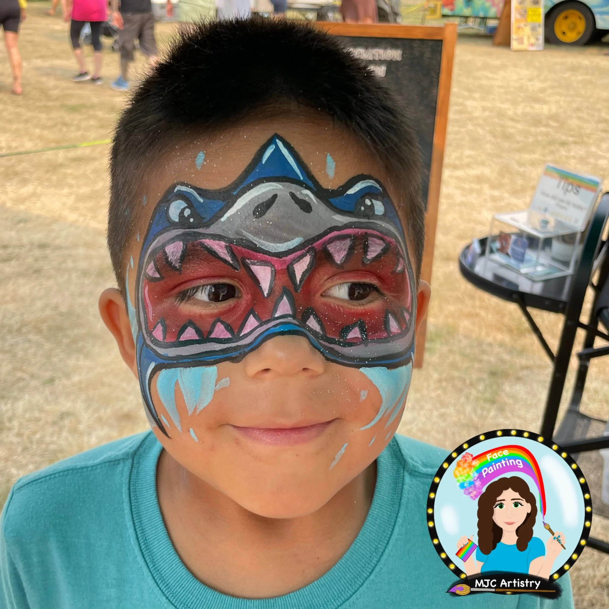 Young boy with colorful face paint resembling a cartoonish dragon or creature covering his eyes and face, outdoors at a fair or festival.