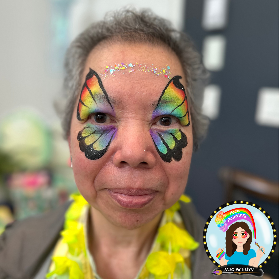 Woman with colorful butterfly face painting, rainbow design, and glitter on forehead, smiling at a birthday party in Vancouver.