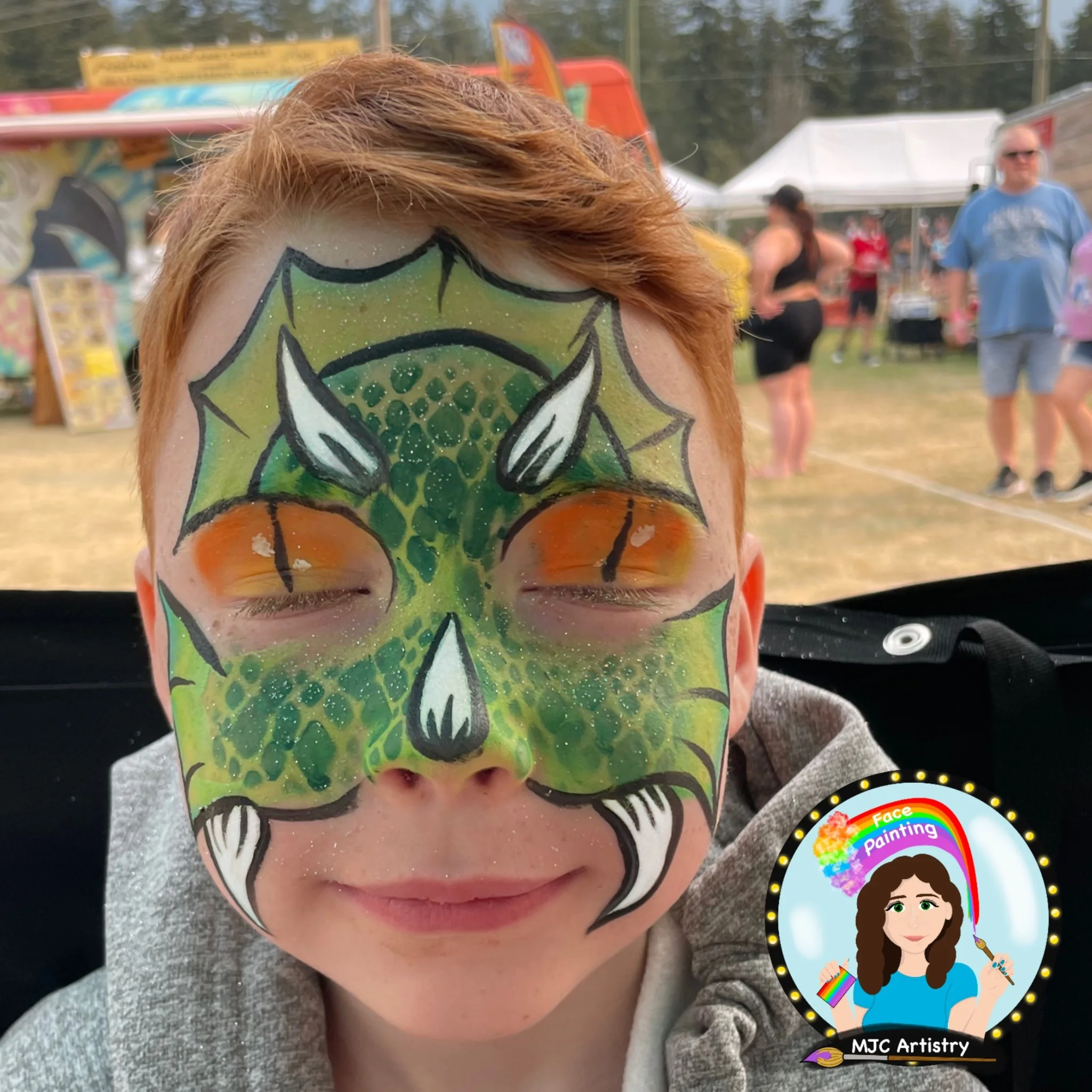Child with colorful face paint resembling a green dragon or dinosaur, with closed eyes, at an outdoor festival or fair with tents and other people in the background at an event in Vancouver BC.