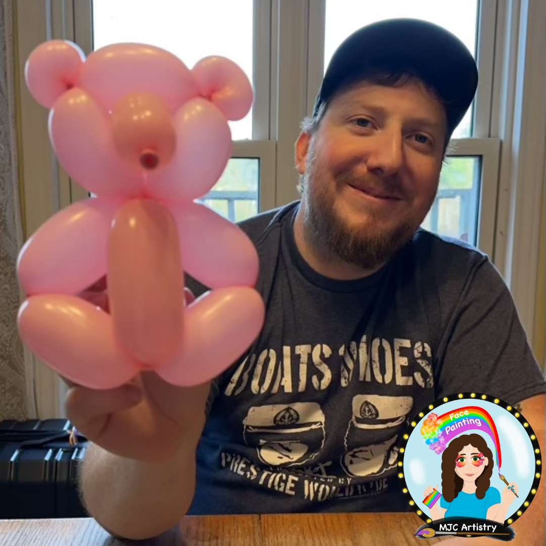 A man with a beard and wearing a black baseball cap holds up a balloon animal shaped like a pink pig, smiling and sitting in front of a window.