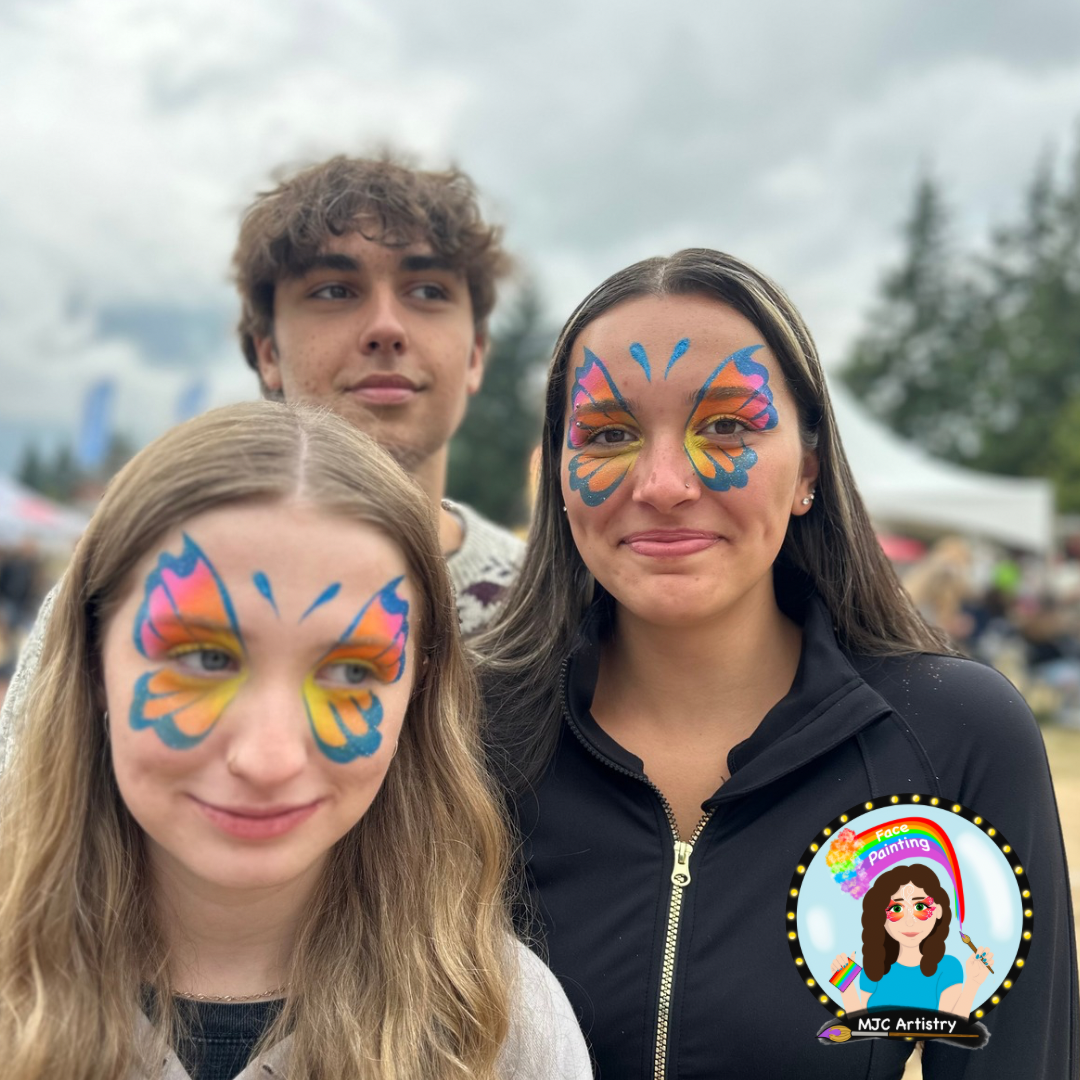 Three people with butterfly face paint, outdoors on cloudy day, background shows tents and trees at an outdoor event in Vancouver.