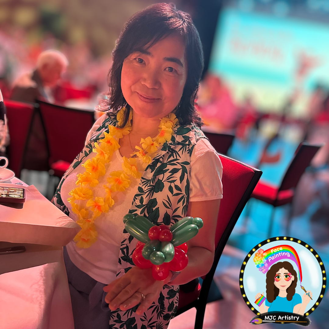 A woman with shoulder-length dark hair sitting at a table in a festive setting. She is wearing a white shirt, a black and white patterned vest, a yellow floral lei, and has a balloon flower in her hand. The background shows other people and a large s