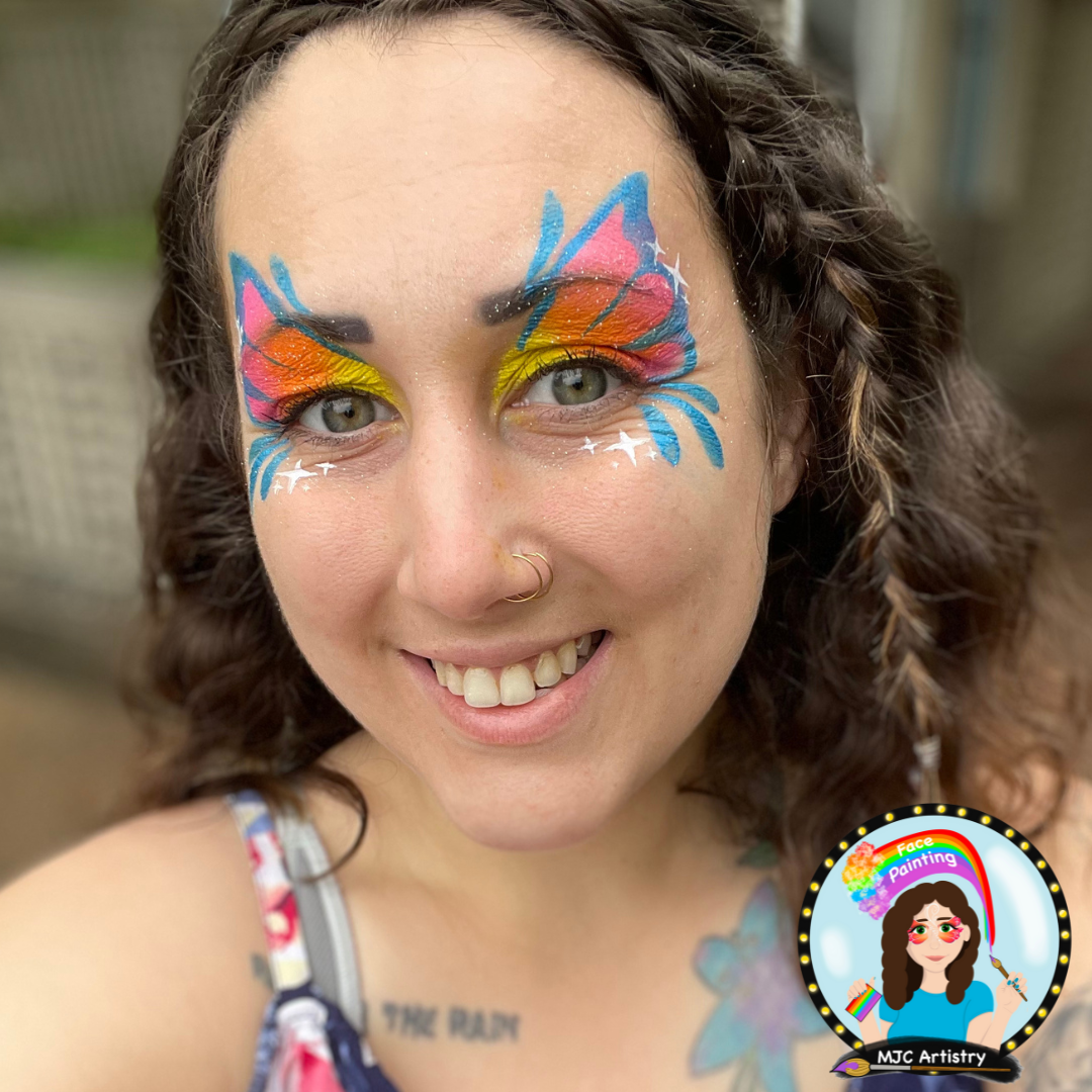 Woman with vibrant rainbow face paint resembling a butterfly around her eyes, smiling at the camera.