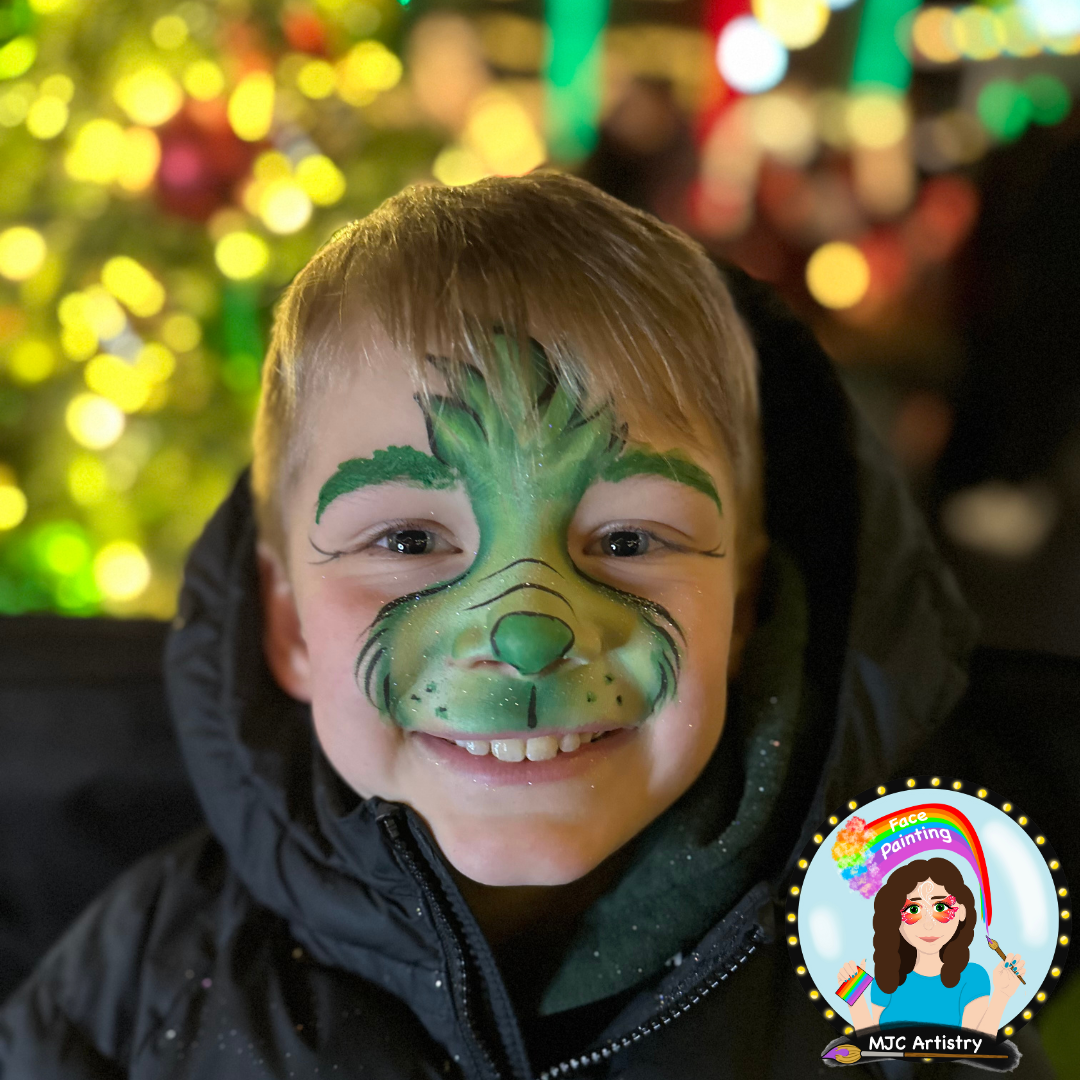 A young boy with a grinch face painting l, smiling in front of blurred colorful Christmas lights at a community event in Vancouver BC. 