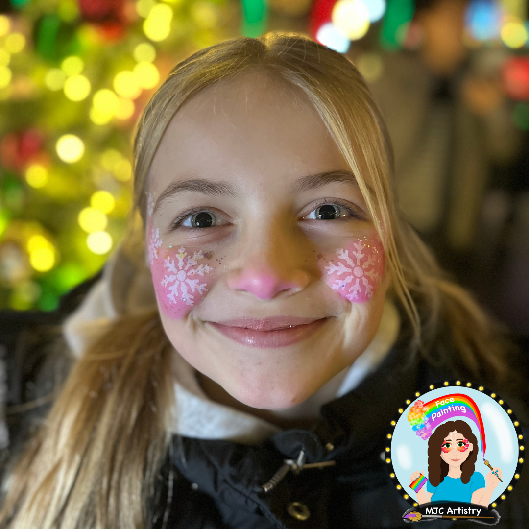 Young girl with face paint of pink snowflakes on her cheeks, smiling, with colorful holiday lights in the background at an event in Vancouver BC. 