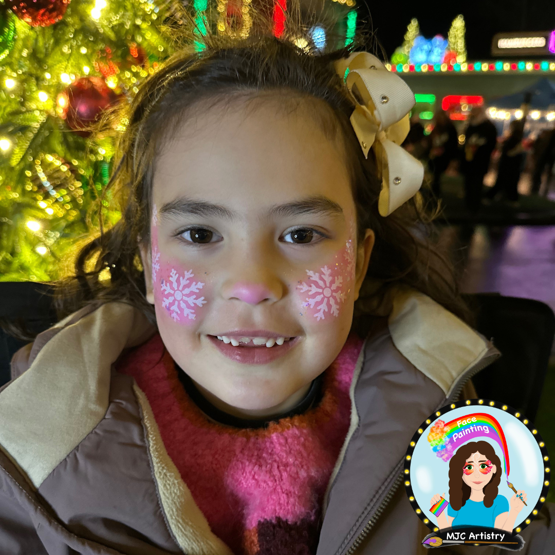 A young girl with face paint of pink snowflakes on her cheeks, smiling, with a decorated Christmas tree in the background at night at a Christmas event in Vancouver BC.
