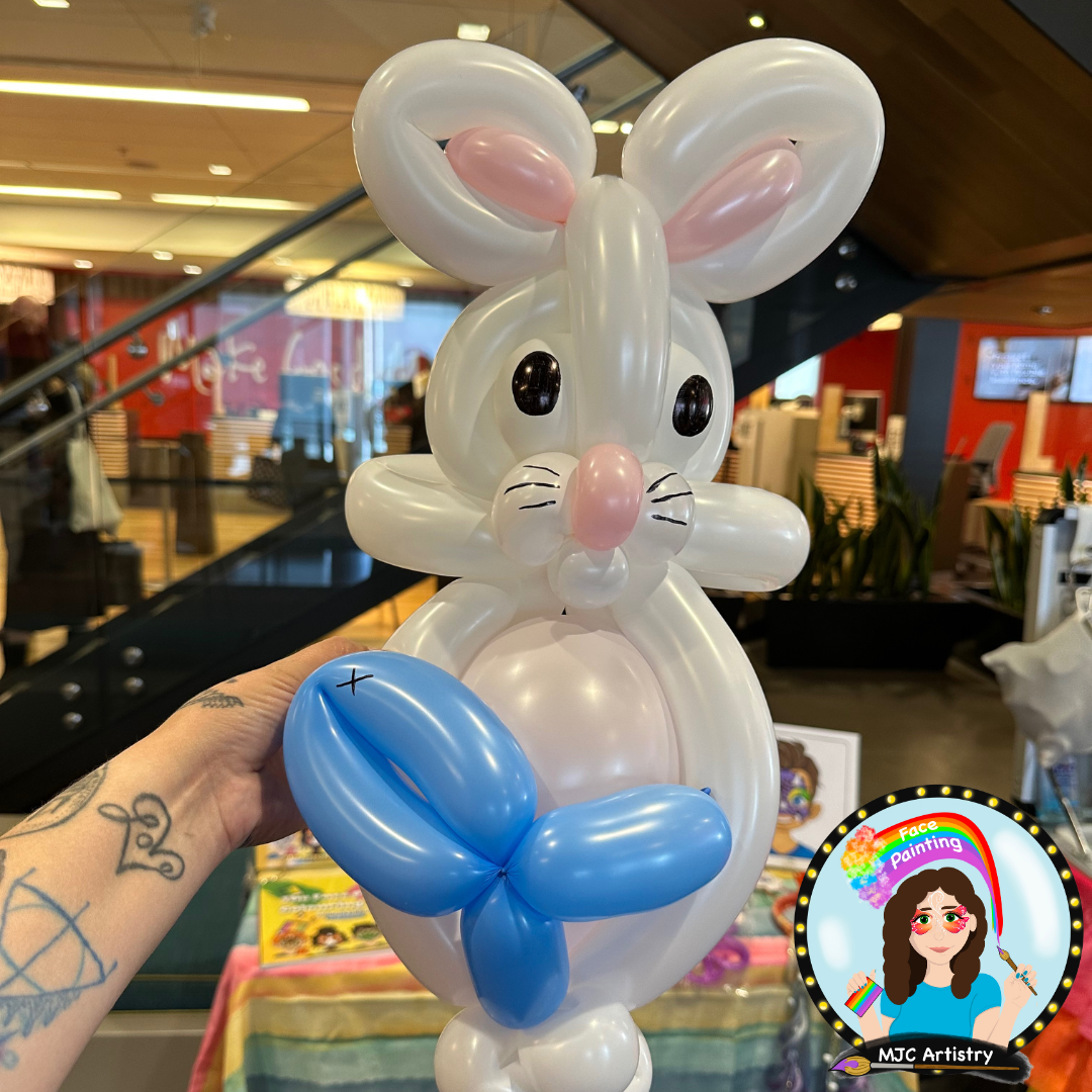 A balloon sculpture of a white bunny with pink ears, a pink nose, black eyes, and a blue butterfly, held by a person with tattoos on their arm, inside a brightly lit indoor space, possibly a store or mall.