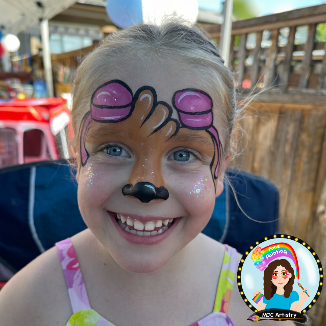 Young girl with face painted to resemble a dog from paw patrol, featuring a black nose, pink and purple accents, and glitter on her cheeks, smiling outdoors at a birthday party in Vancouver BC.