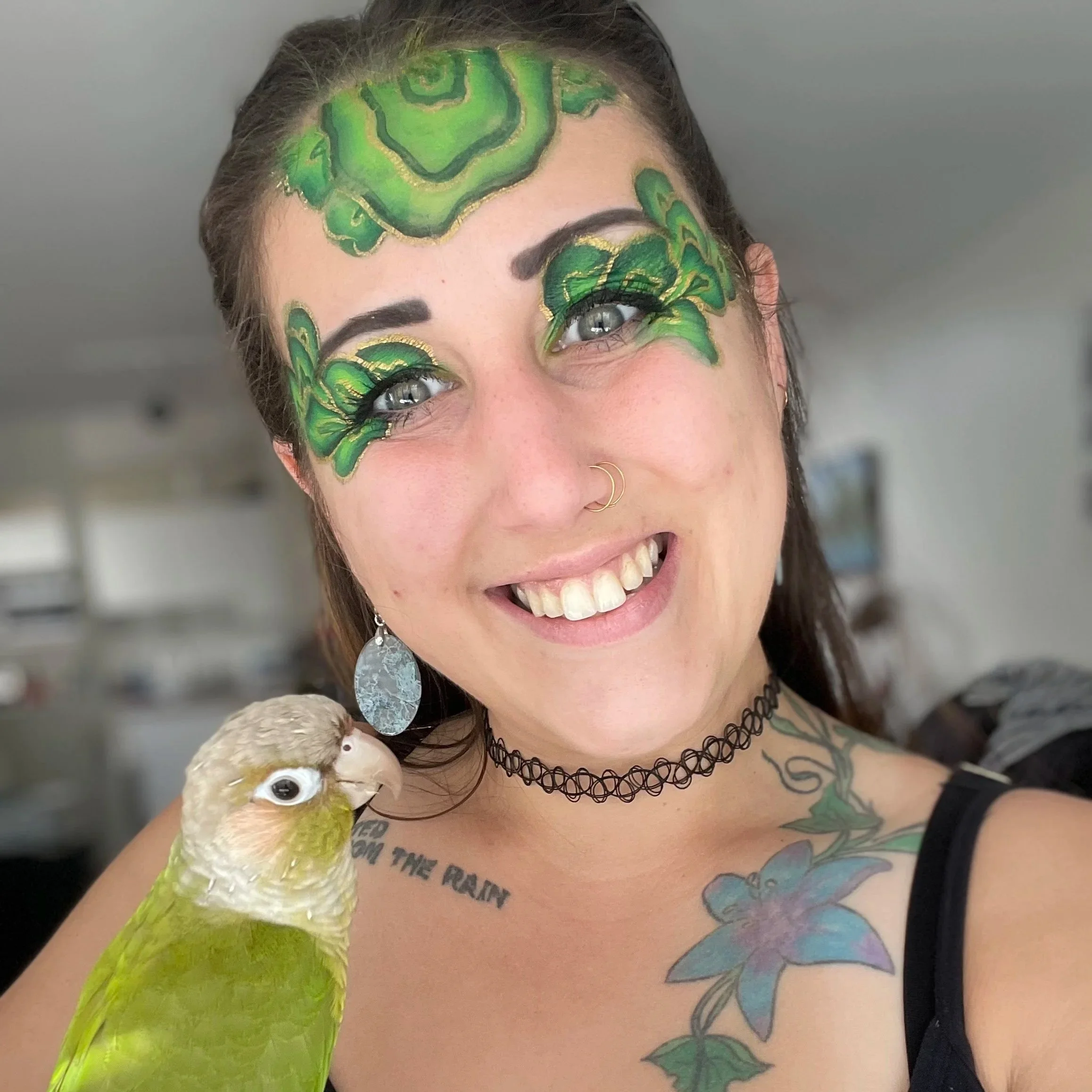 A woman with face makeup resembling green leaves, a tattoo on her chest, a black choker, and earrings, smiling while holding a green and white bird.
