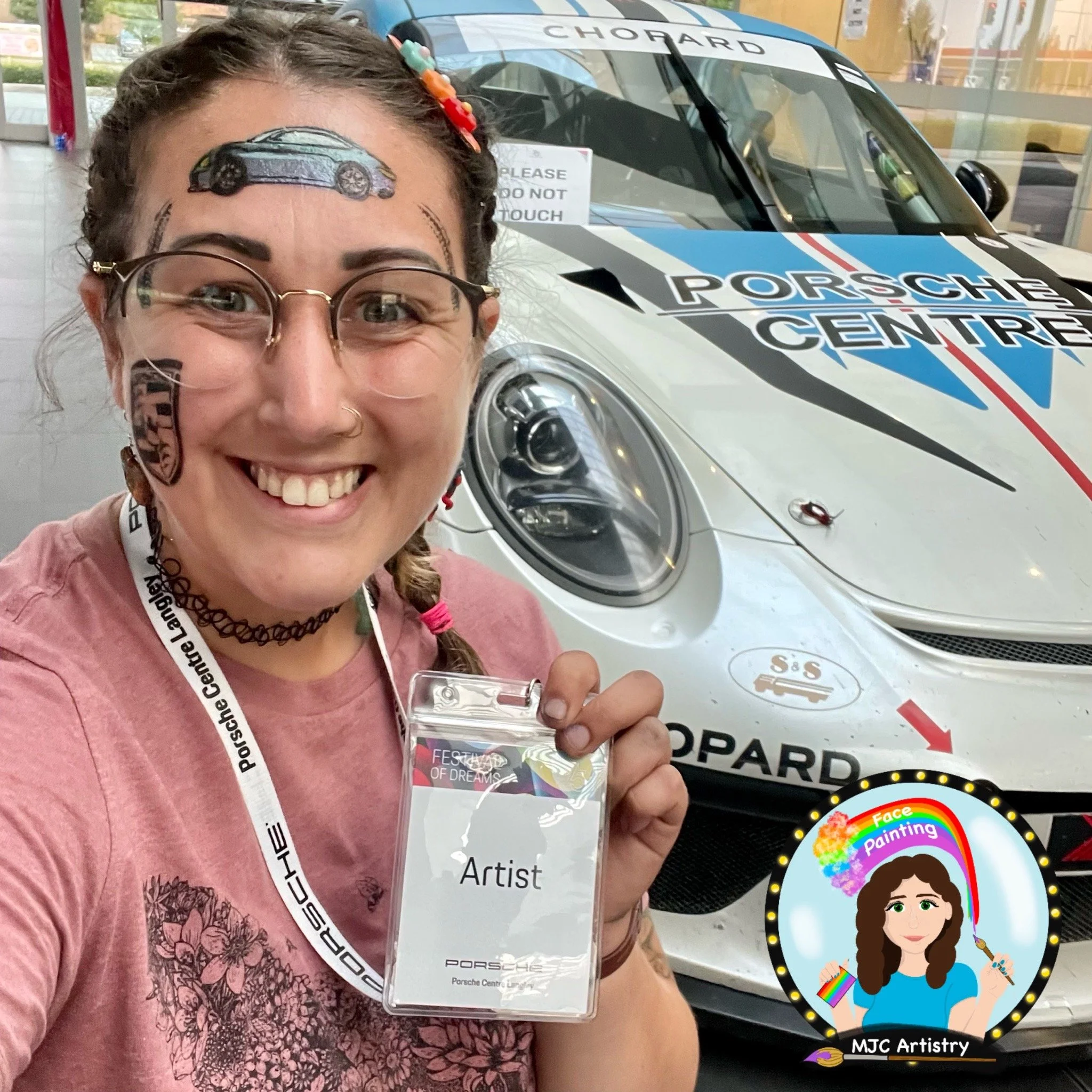 A woman with glasses, braided hair with colorful beads, and a nose ring smiling and holding an 'Artist' badge at a Porsche event. Behind her is a white Porsche race car with Porsche Center branding.