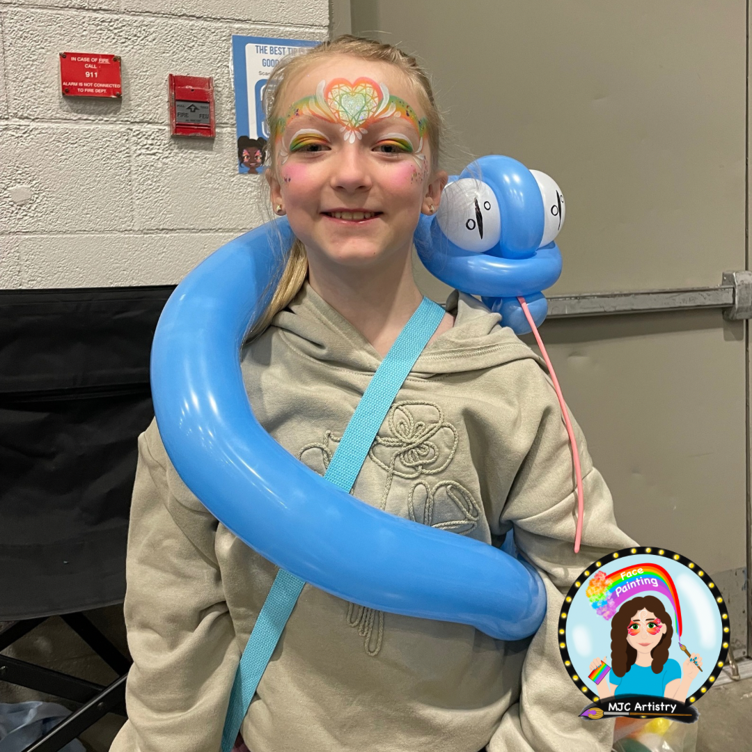 A young girl with rainbow face paint, featuring a colorful heart-shaped design on her forehead and pink cheeks, smiling at the camera. She is wearing a beige hoodie and has a blue balloon twisted around her neck and shoulder, with a balloon sculpture