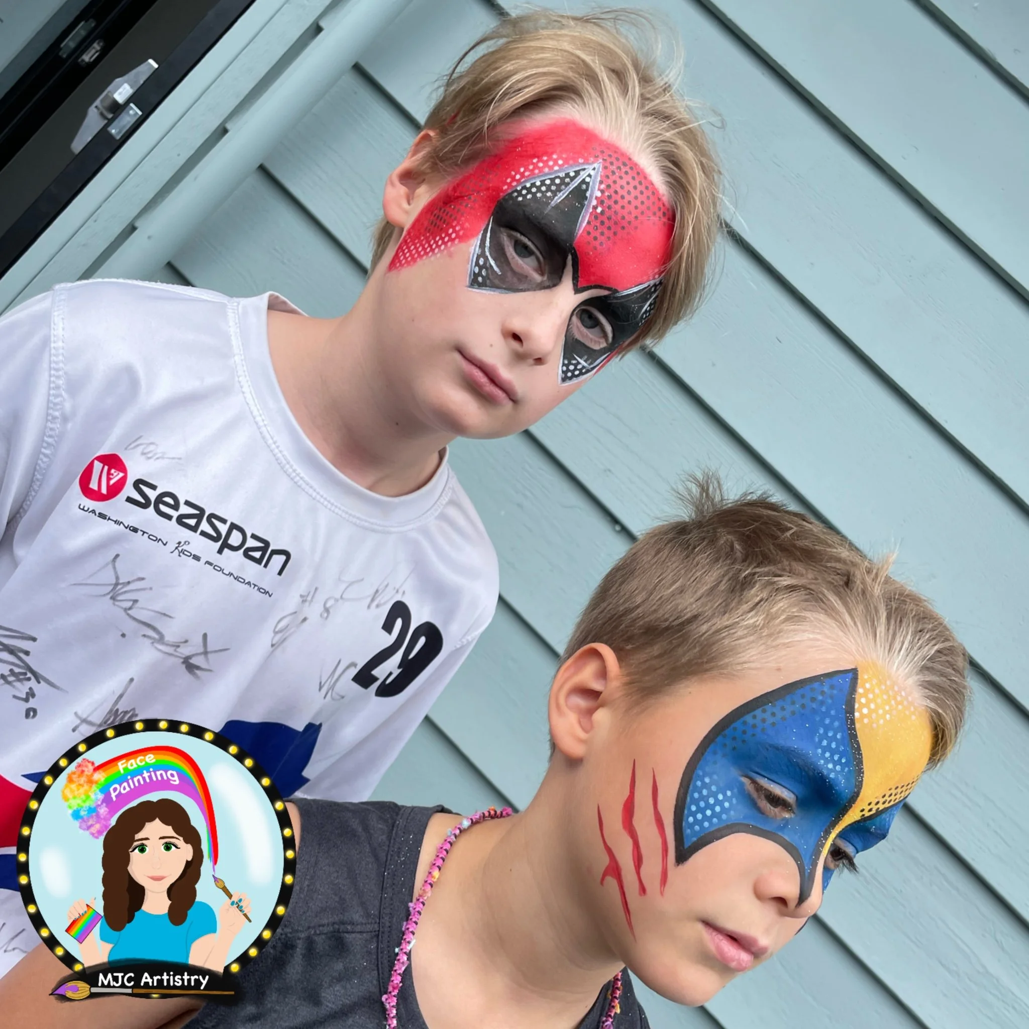 Two children with face paint standing in front of a light blue wooden wall. The taller child has a red and black face design resembling a superhero mask, while the shorter child's face is painted with blue and yellow colors, with red claw marks on th