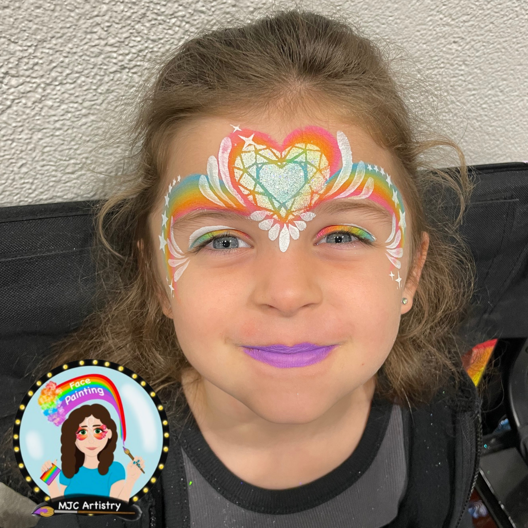 Young girl with rainbow and heart face painting on her forehead, wearing purple lipstick and smiling at a birthday party in Vancouver.