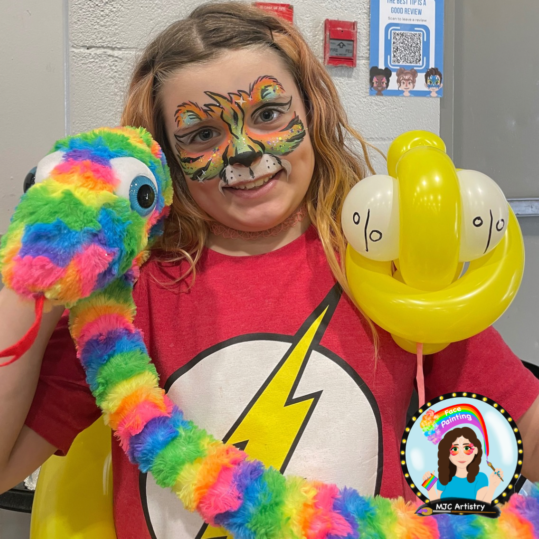 A young girl with face paint resembling a tiger, holding a rainbow-colored plush chameleon and yellow balloon sculpture shaped like a smiling face with balloons as cheeks, smiling at the camera.