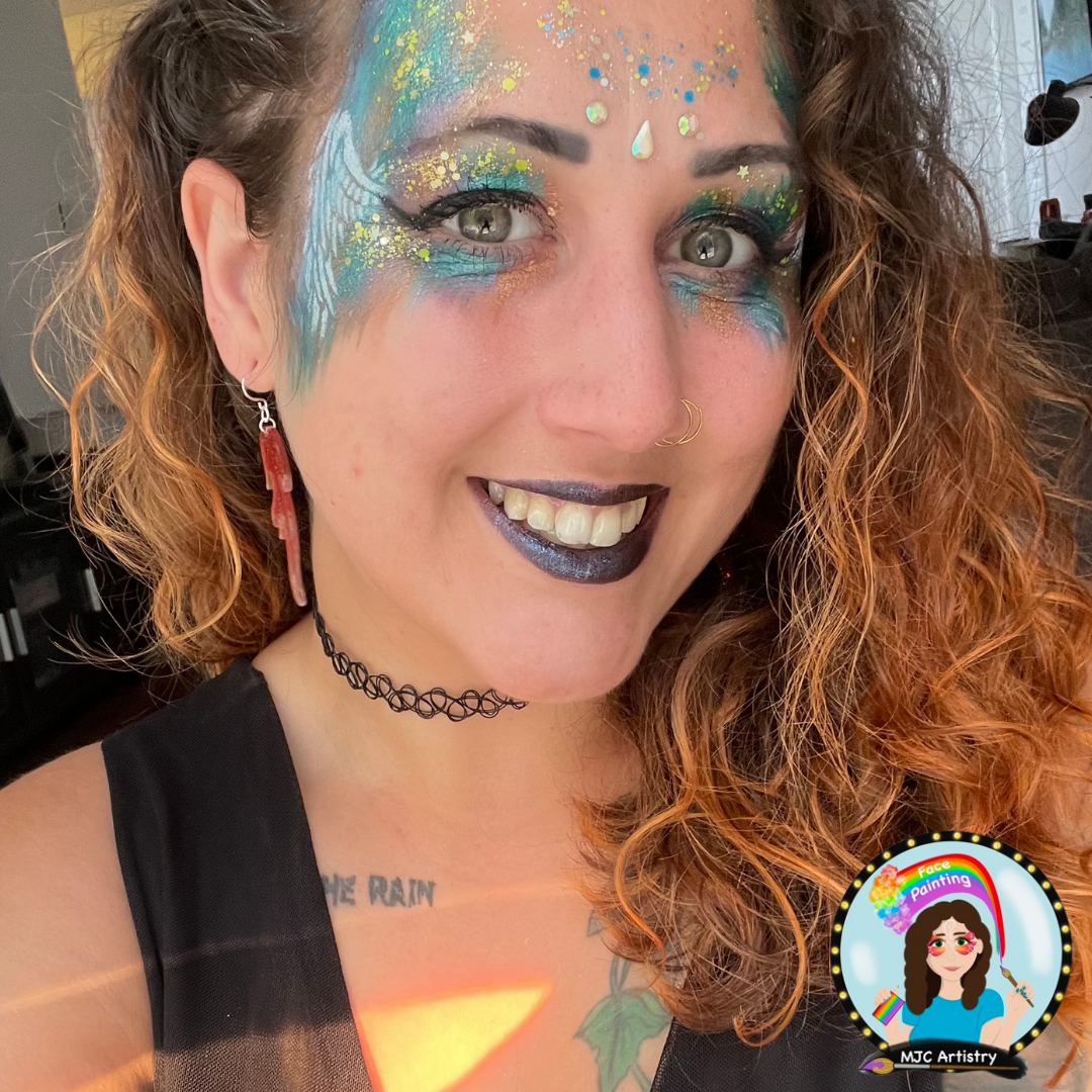 Woman with curly red hair wearing vibrant rainbow face paint, glitter, and makeup, smiling, with a tattoo on her chest and a black choker necklace. She is outdoors with a background that appears to be a room or studio.