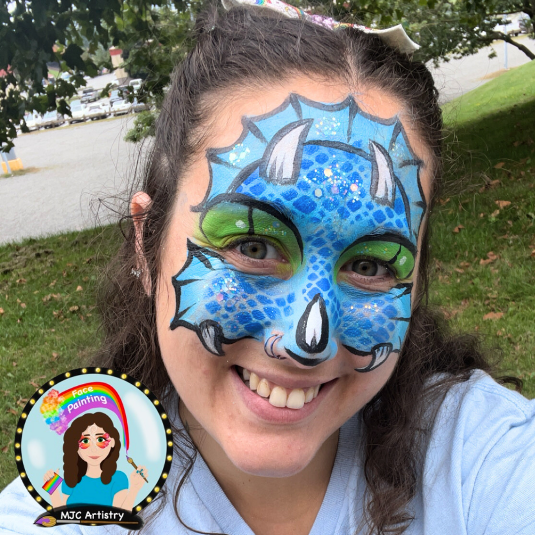A woman smiling outdoors with colorful face paint that resembles a blue dragon with green eyeshadow and black outlines.