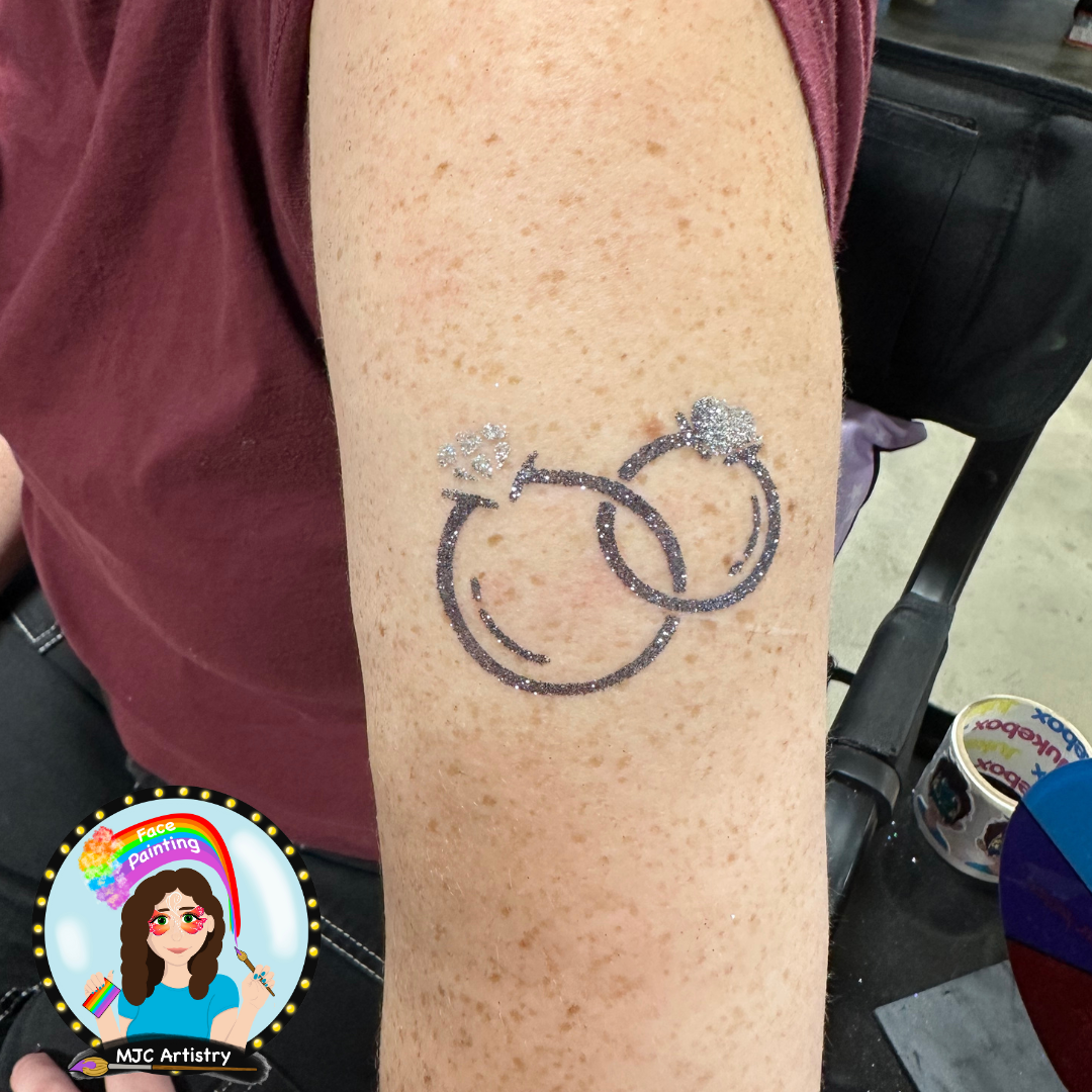 A temporary tattoo of two intertwined wedding rings with small hearts and glitter on a person's arm.
