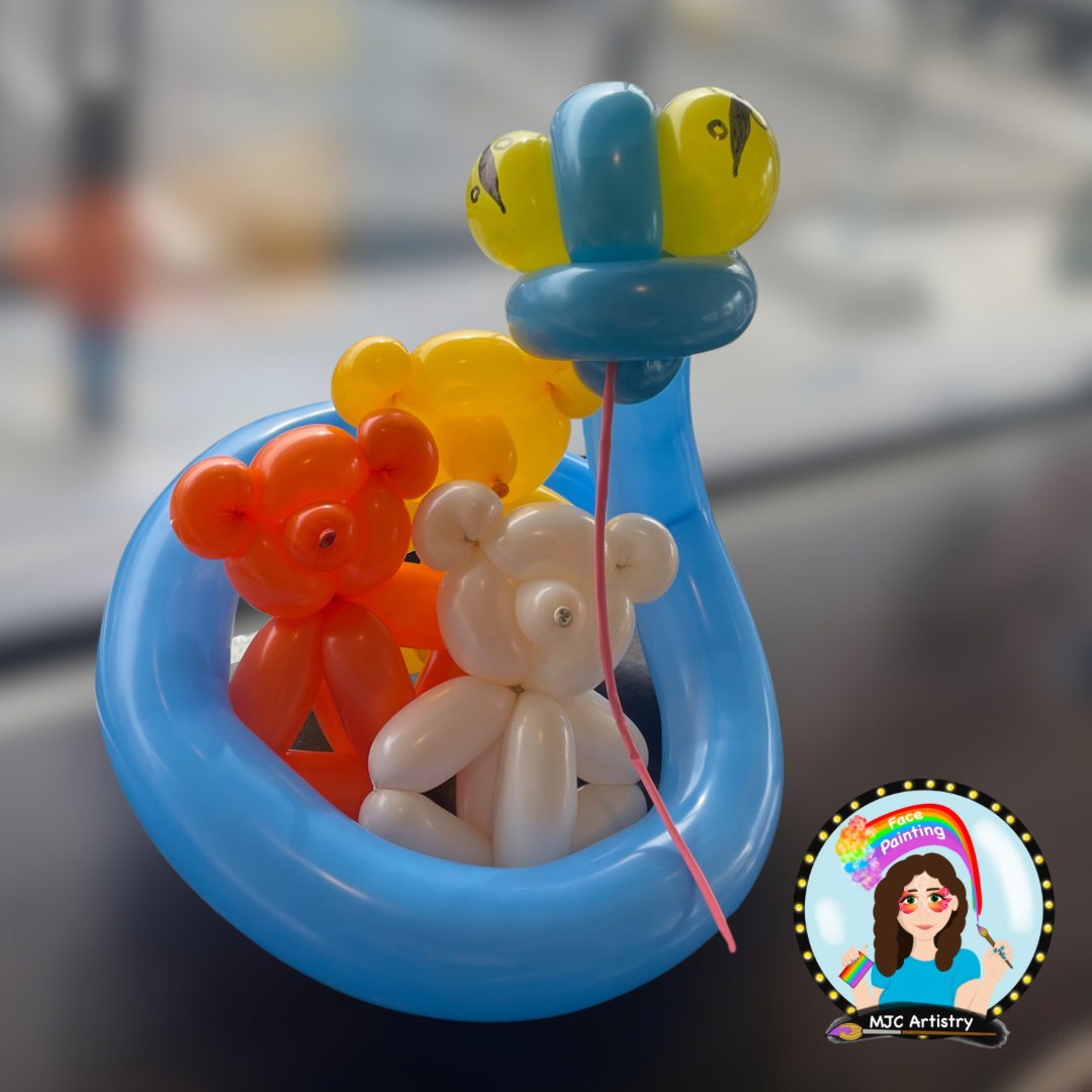 Colorful balloon sculpture of a bear with a rainbow-colored face, surrounded by other balloons, inside a blue balloon basket, with a logo in the bottom right corner featuring a girl holding a paintbrush and a rainbow.