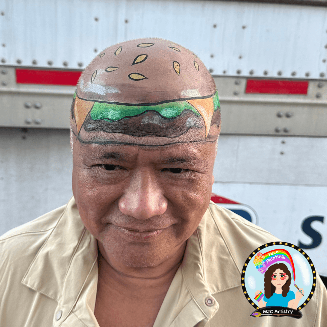 A man wearing a beige shirt with a painted design on his bald head resembling a hamburger, with sesame seed bun, lettuce, tomato, cheese, and beef patty.
