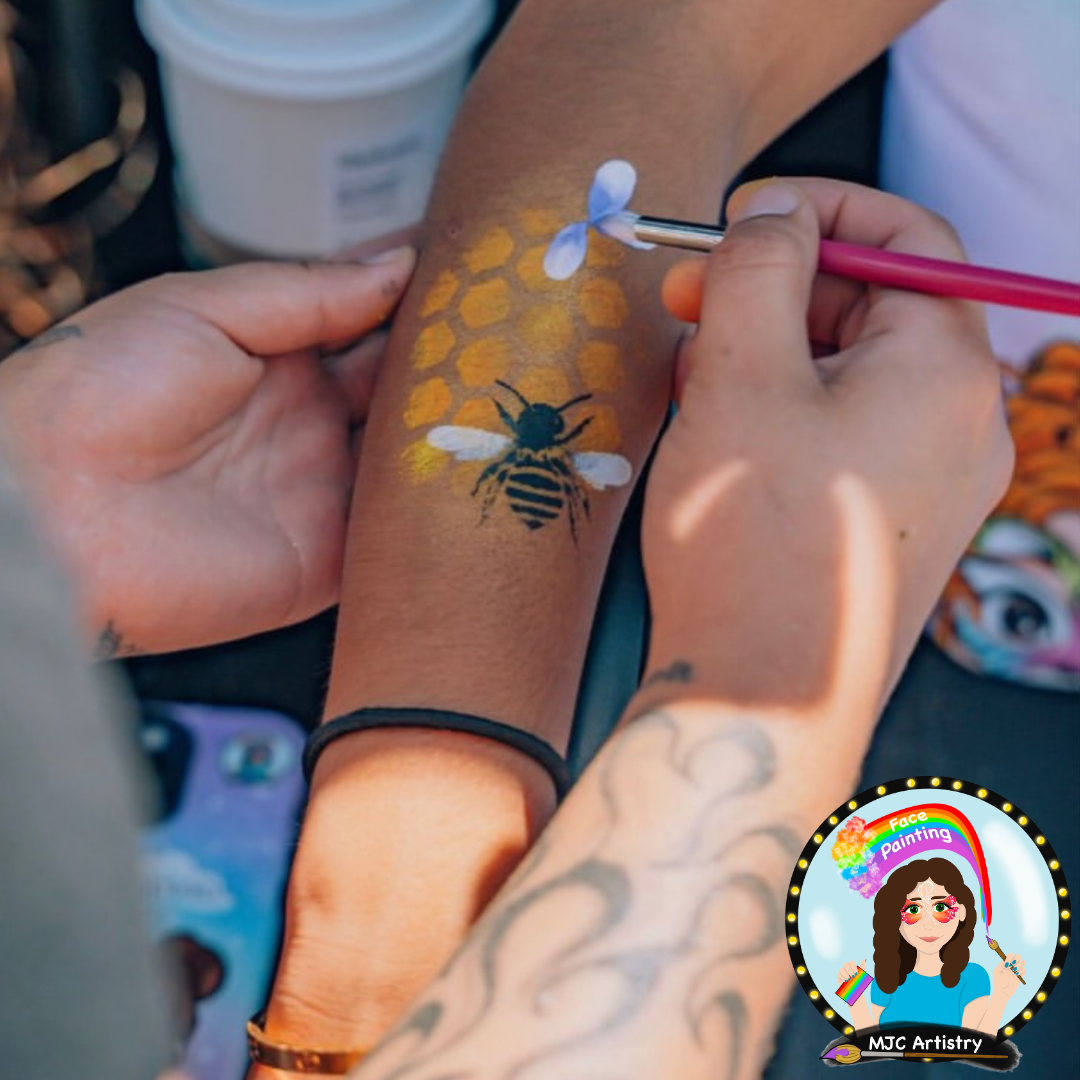 Close-up of a person receiving face painting on their arm, depicting a bee and honeycomb design. The artist is using a pink brush to add details. The person has tattoos on their other arm and wears a black hair elastic. A cup of drink and other items