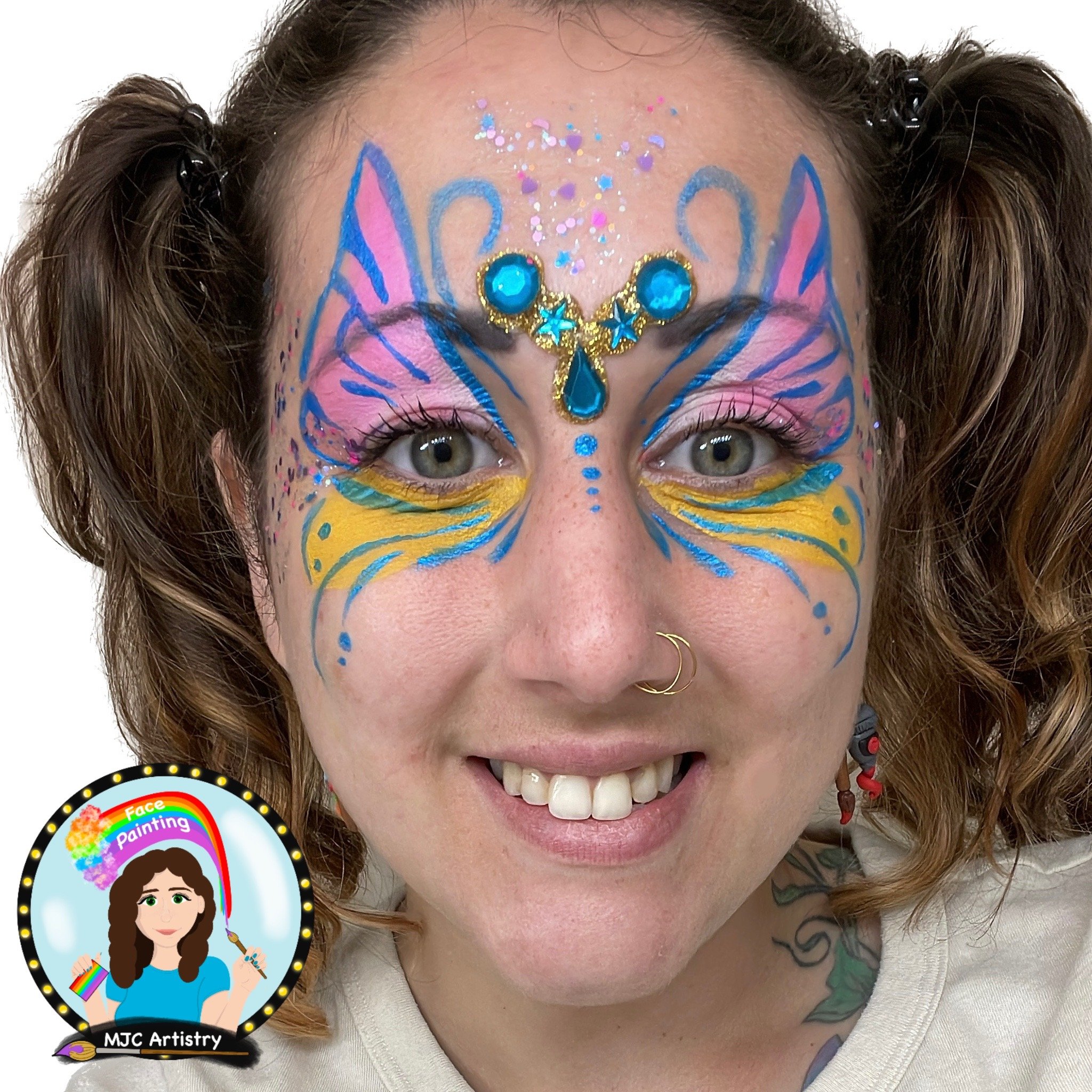 Woman with face painted as a colorful butterfly, featuring pink, blue, and yellow designs, blue gemstone forehead decoration, and a nose ring, smiling at the camera.