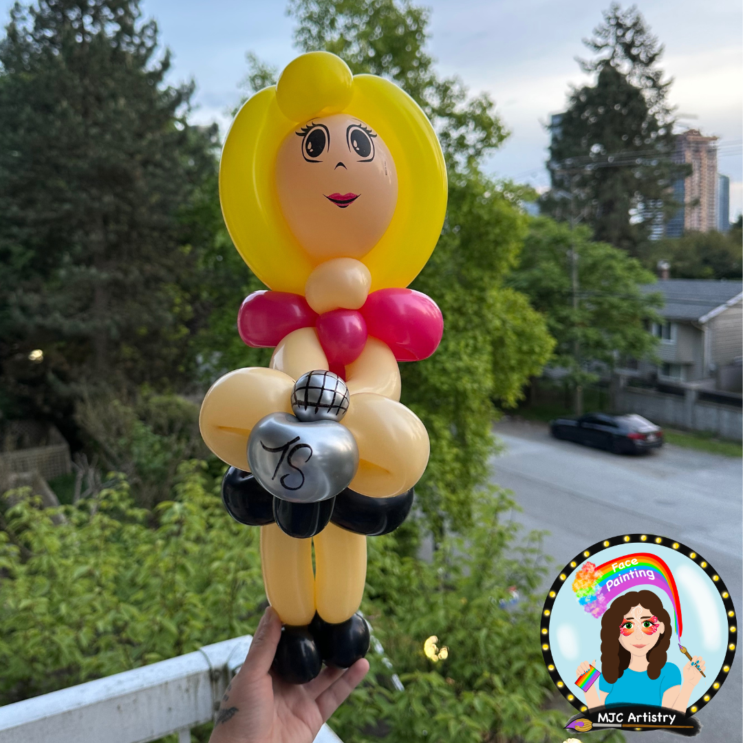 Balloon sculpture of a woman with blonde hair, red bow, and black shoes, holding a silver balloon with the number 15. The sculpture has a cartoon face with big eyes and red lips.