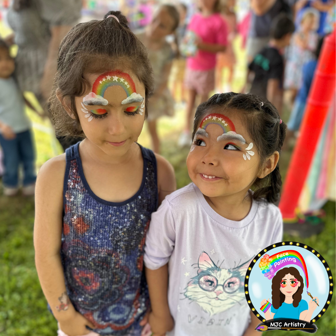 Two young girls with rainbow and cloud face paint, standing outdoors at a festival or fair, surrounded by other children and adults.