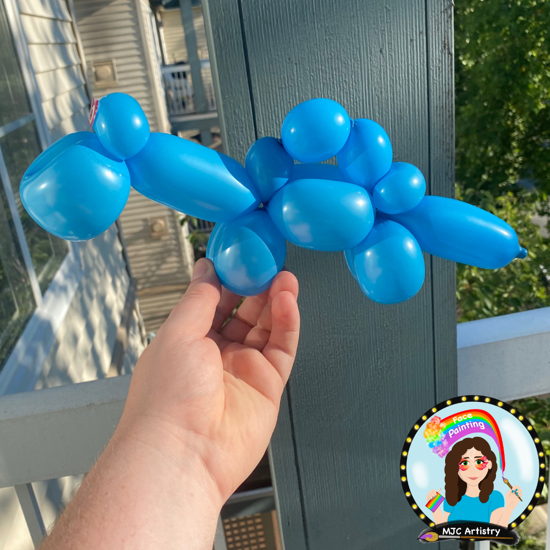 Blue balloon sculpture resembling a puppy held outdoors near a blue wooden post with trees in the background.