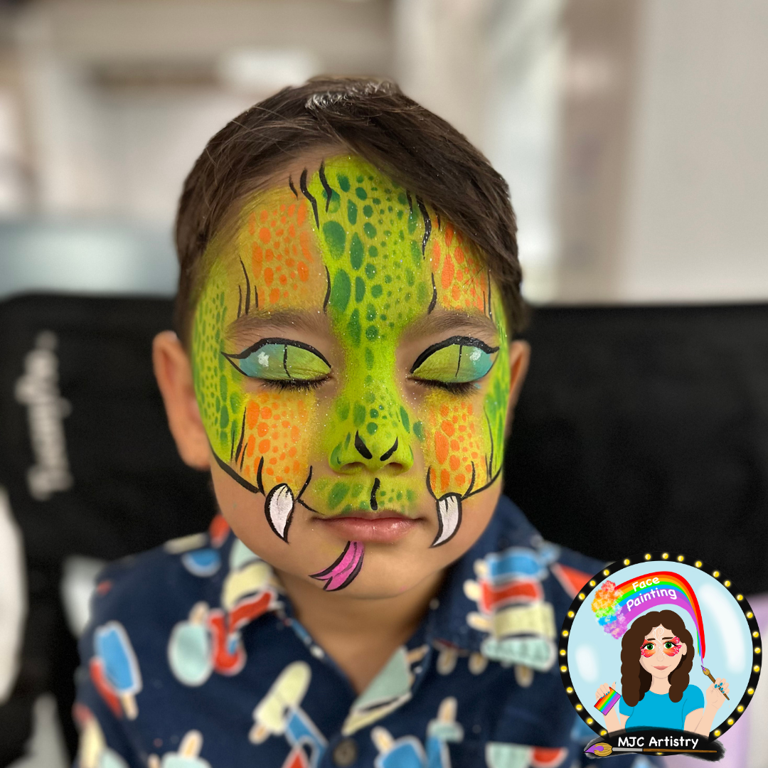Child with face painted as a colorful snake, featuring green, yellow, orange, and black details, horns, and a tongue sticking out, wearing a blue shirt with ice cream patterns at a birthday party in Vancouver. 