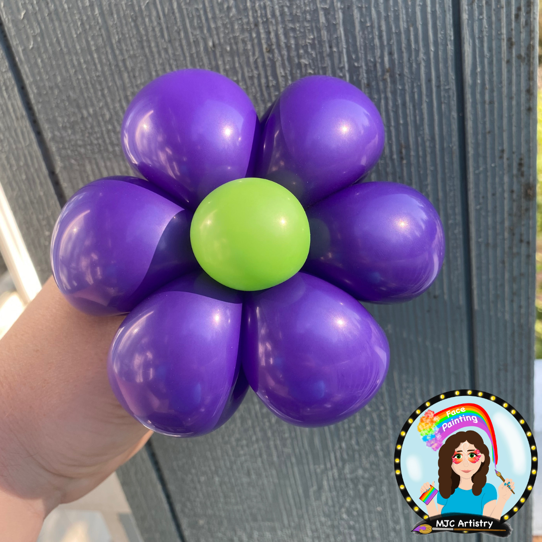A hand holding a balloon flower with purple petals and a green center, set against a wooden wall.
