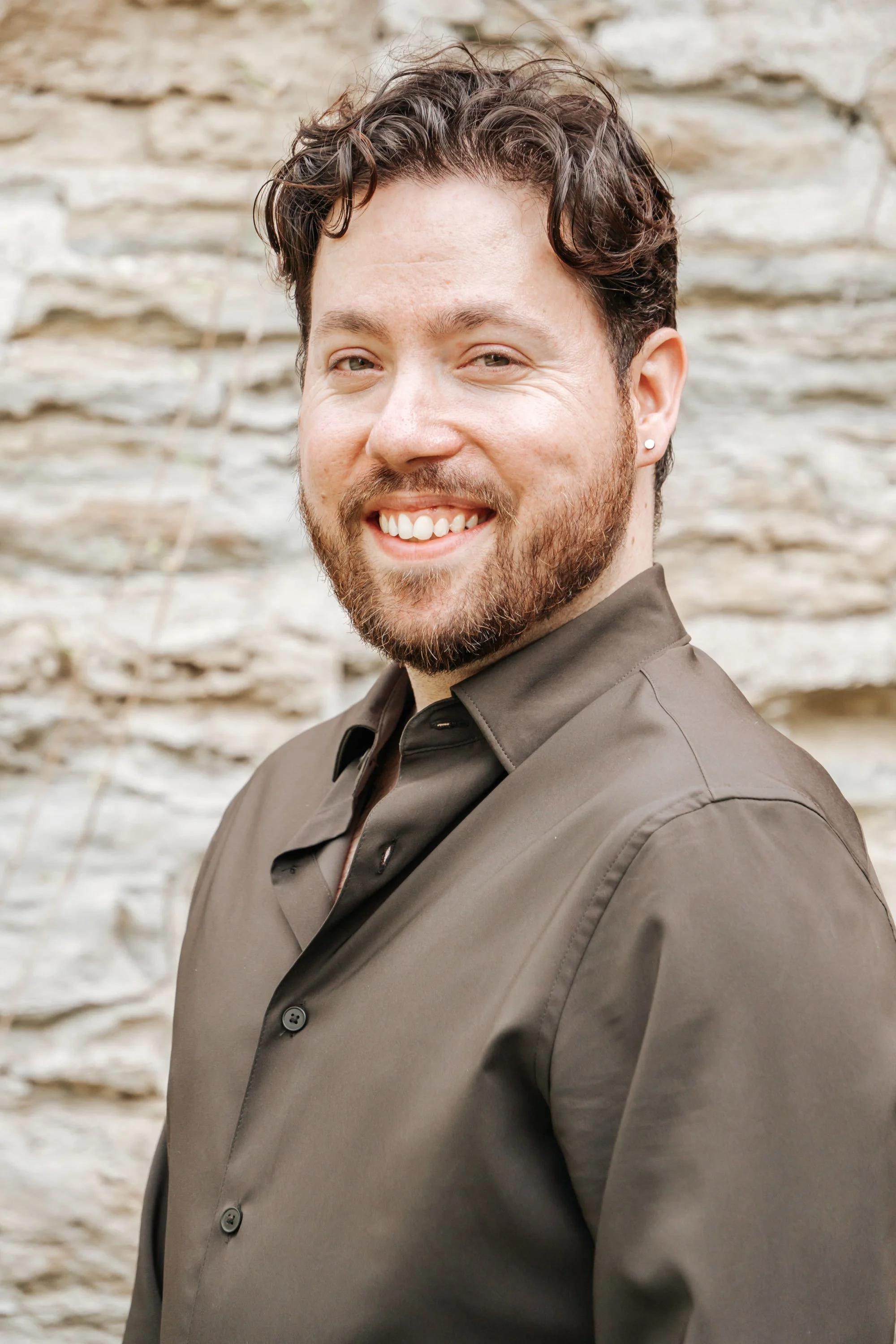 A man with curly brown hair and beard, wearing a dark gray shirt, smiling, standing against a stone wall background.