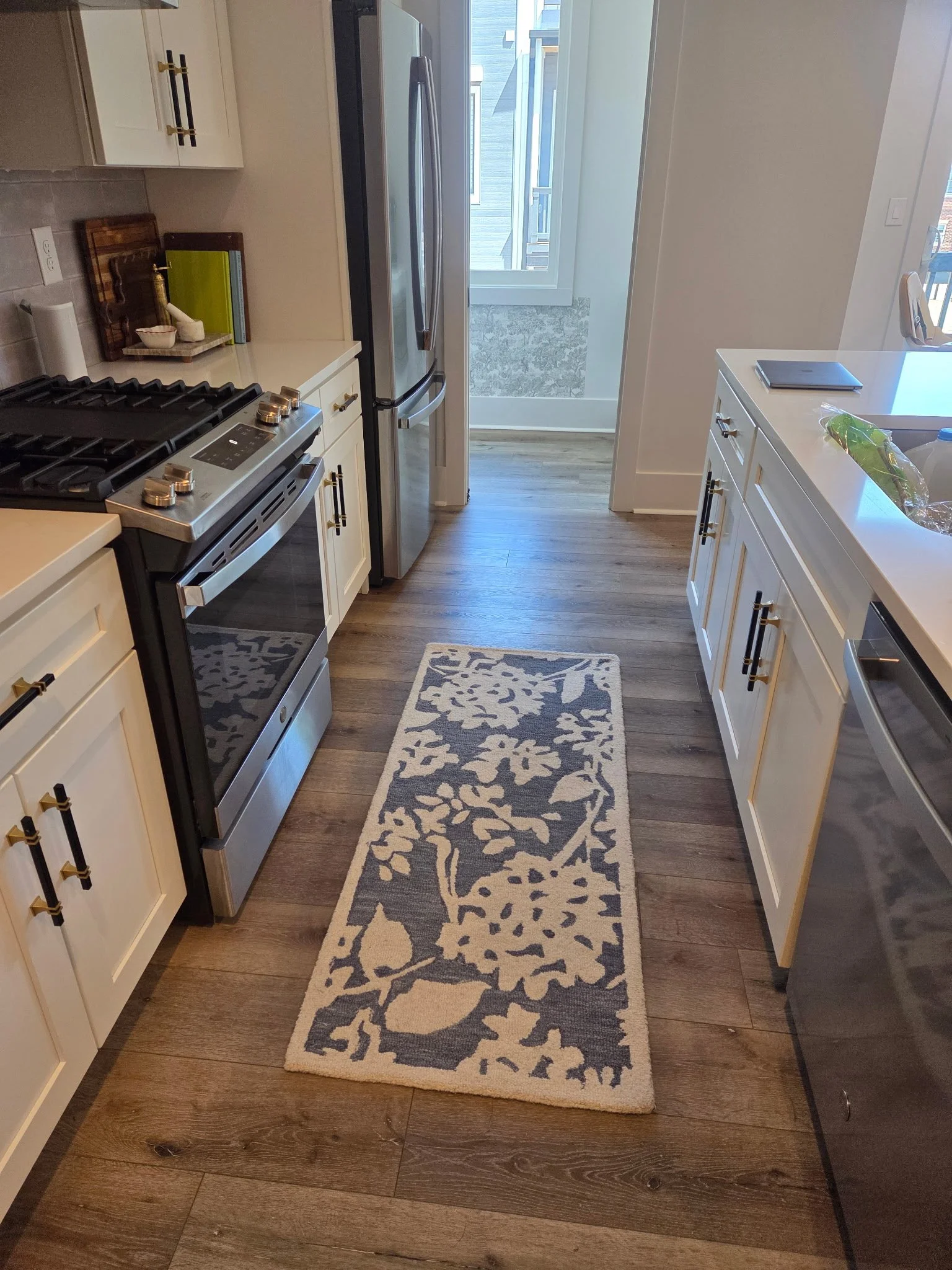 A kitchen with white cabinets, a stainless steel refrigerator, a gray and white patterned rug on brown wood flooring, and a white countertop with items including a laptop, a plastic bag containing green vegetables, and a book. There is a door at the end of the room leading to a balcony or outdoor space with a mountain view.