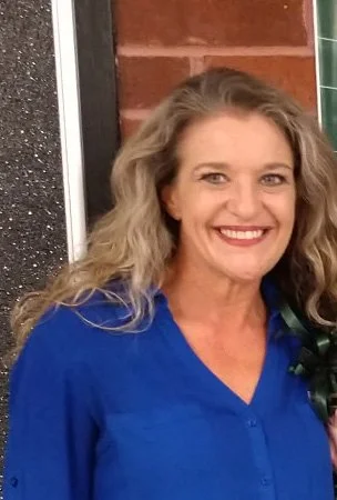 Nikki, the owner, smiling with long wavy blonde hair wearing a blue top, standing indoors.